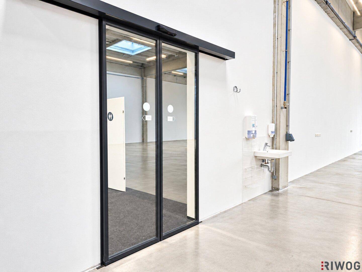 Heller Flurbereich mit automatischen Glastüren, Waschbecken und Seifenspendern an der Wand.