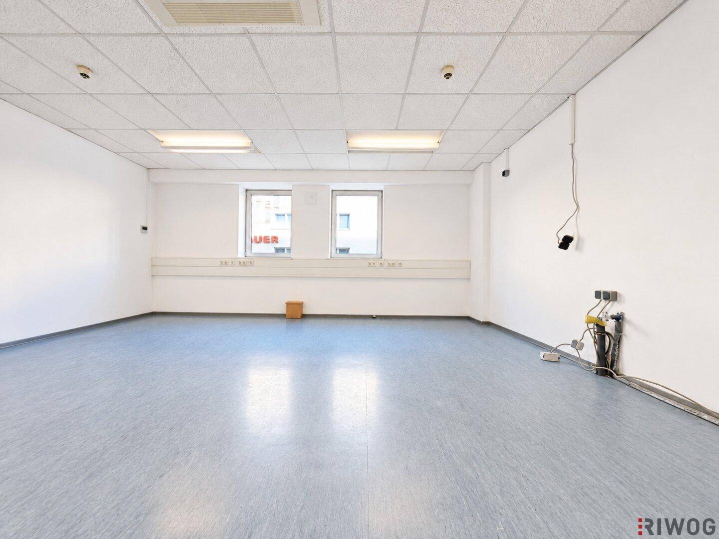 Büroraum mit grauem Linoleumboden, zwei Fenstern und sichtbaren Kabelinstallationen.