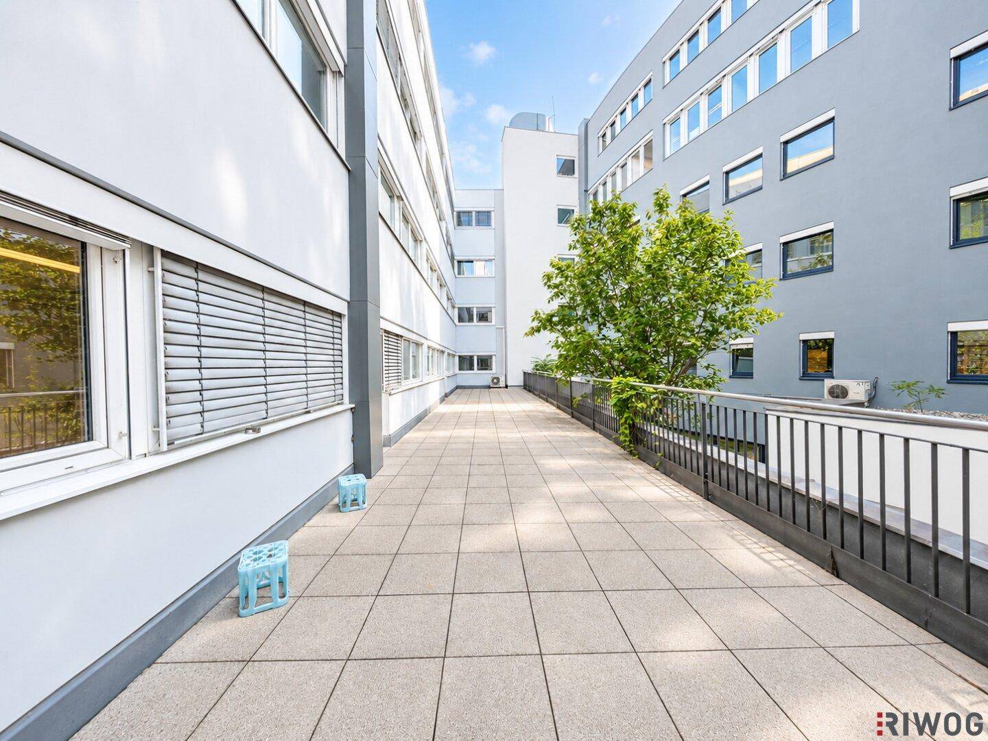 Weitläufige Außenterrasse mit Fliesenboden, Metallgeländer und Blick auf angrenzende Gebäude.