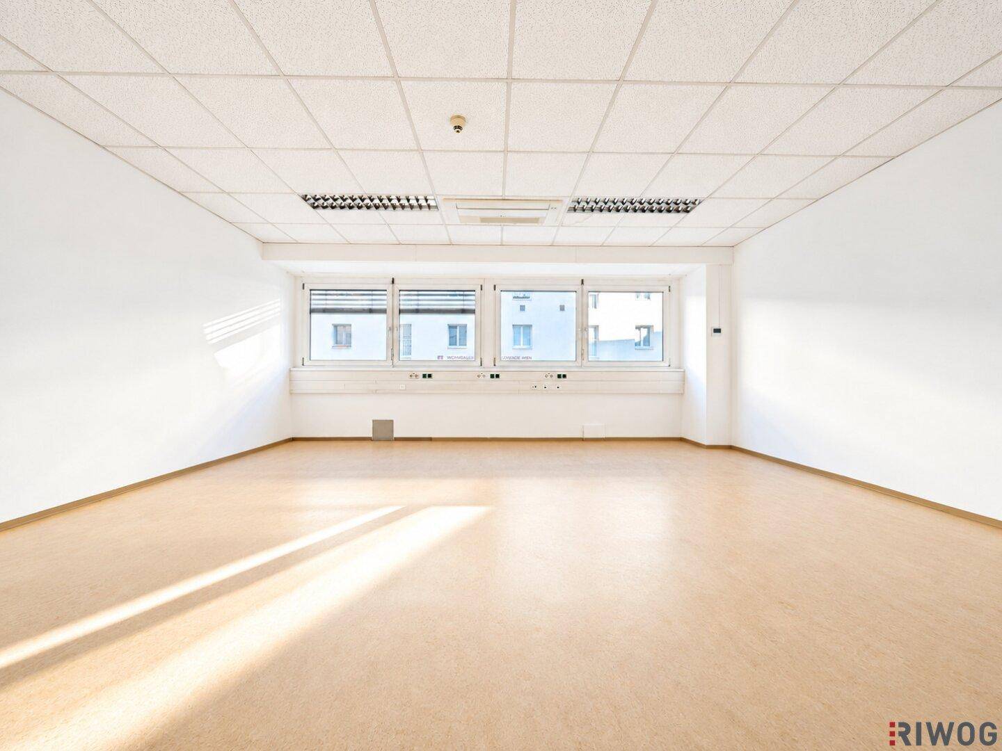 Heller Büroraum mit großer Fensterfront, hellem Boden und Rasterdeckenbeleuchtung.