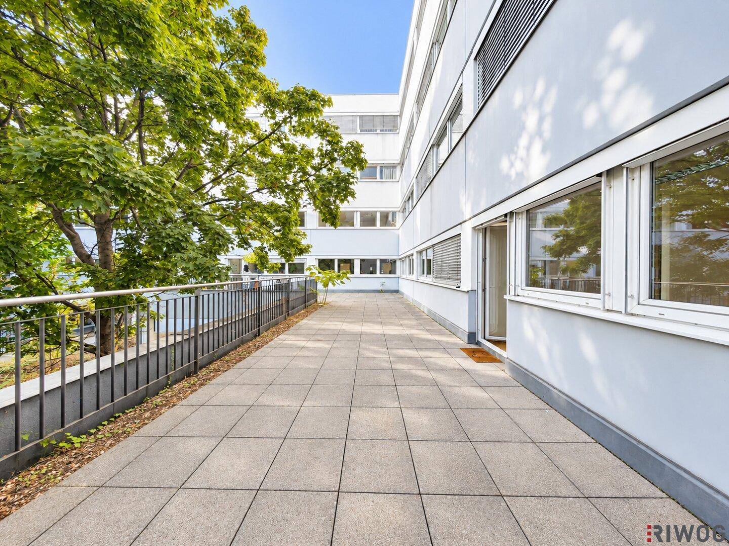 Große Außenterrasse mit Fliesenboden und üppigem Grün, umgeben von Bürogebäuden.