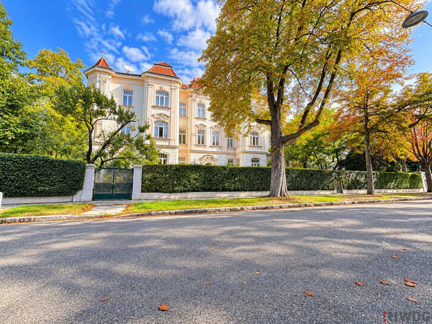 Prächtiges Gebäude mit klassischer Architektur, umgeben von Herbstbäumen und einer grünen Hecke.