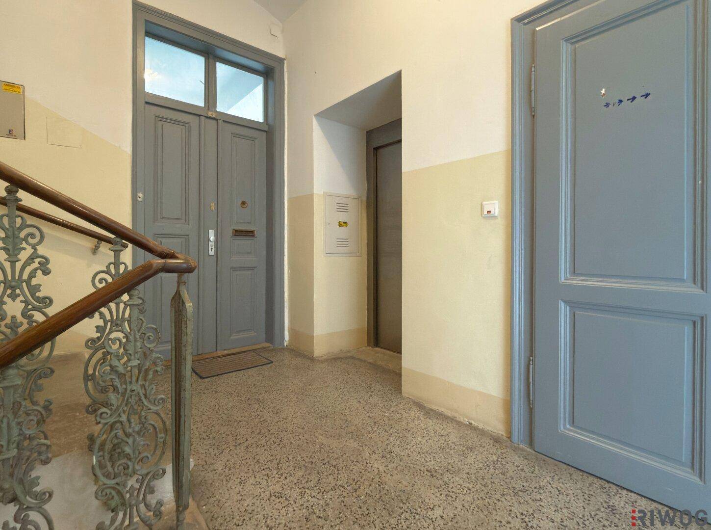 Treppenhaus mit Aufzug, zwei Wohnungstüren und einem historischen Treppengeländer.
