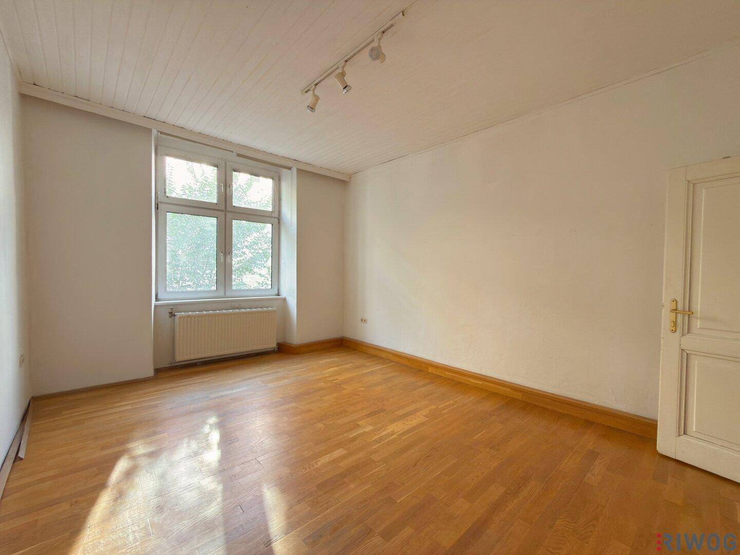 Geräumiger Raum mit Parkettboden, großem Fenster und Heizkörper unter dem Fenster, gut beleuchtet.