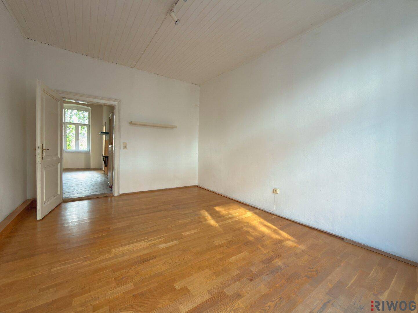 Heller, leerer Raum mit Holzparkettboden, weißen Wänden und einer Tür zu einem weiteren Zimmer.