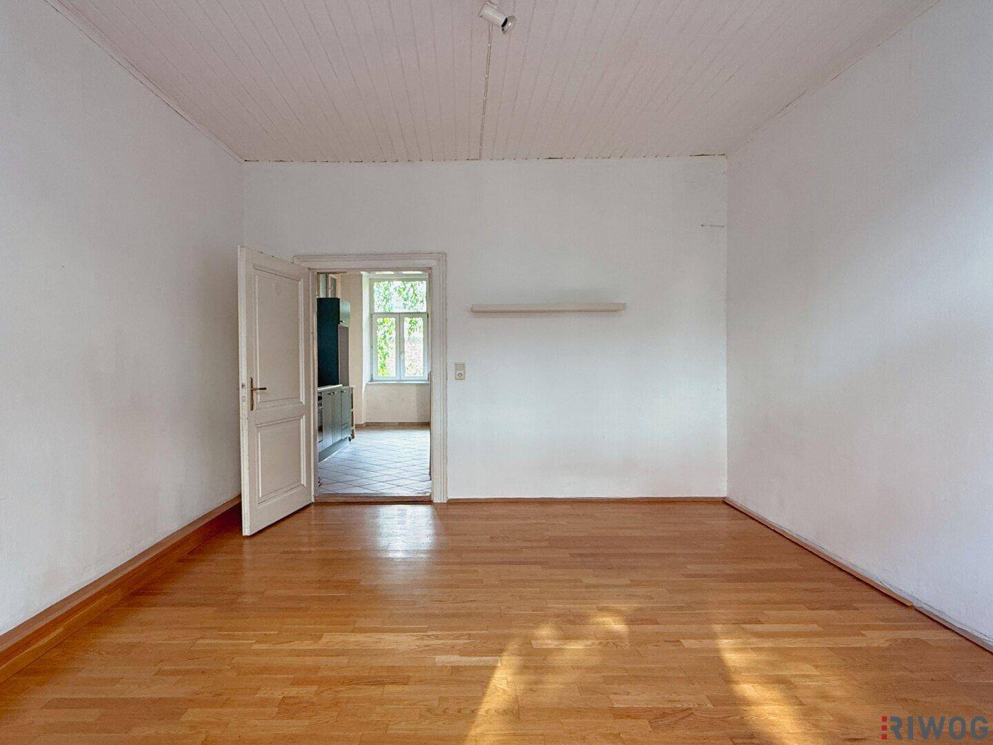 Heller Raum mit Parkettboden und offener Tür zu einem weiteren Zimmer mit Fliesenboden.