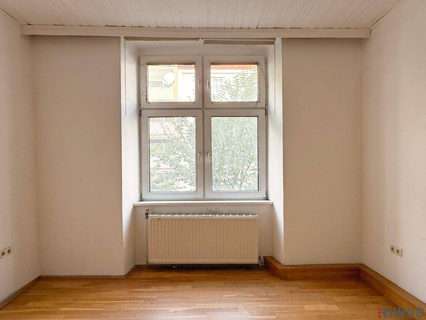 Einfacher Raum mit Parkettboden, weiß gestrichenen Wänden und einem Fenster mit Blick ins Grüne.