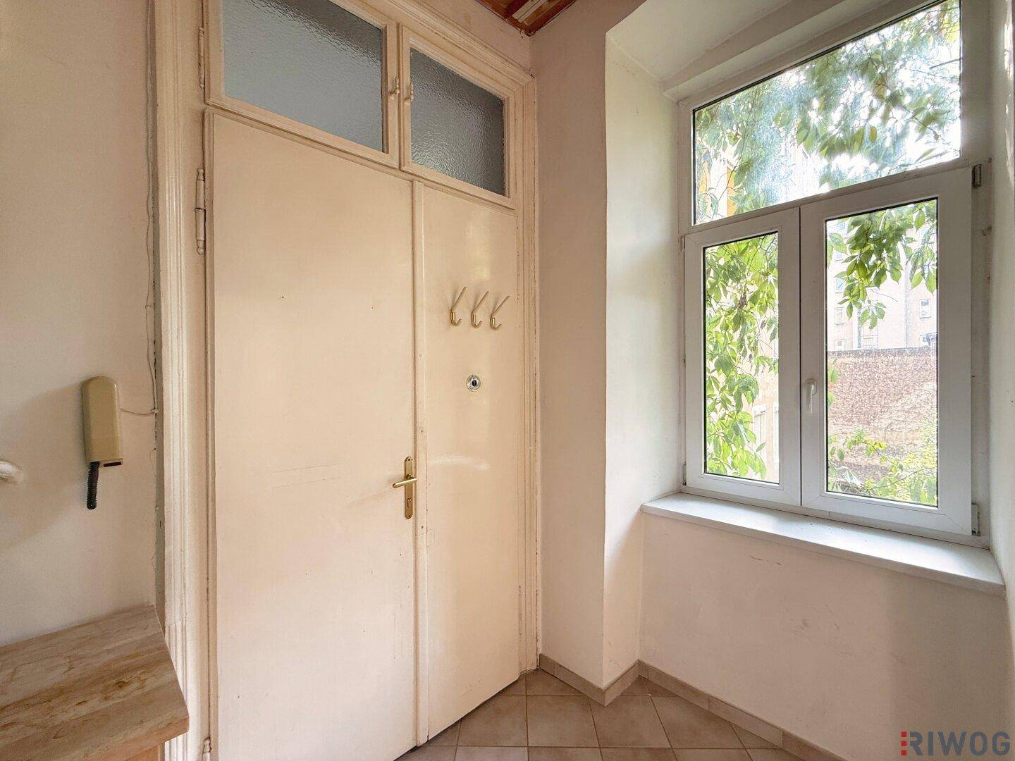 Eingangsbereich mit einer alten Holztür, Fliesenboden und einem Fenster mit Blick ins Grüne.
