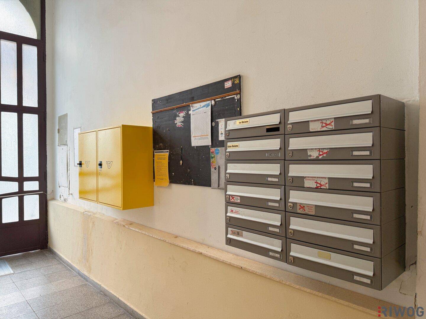 Eingangsbereich eines Mehrfamilienhauses mit Briefkästen, schwarzem Brett und Fliesenboden.