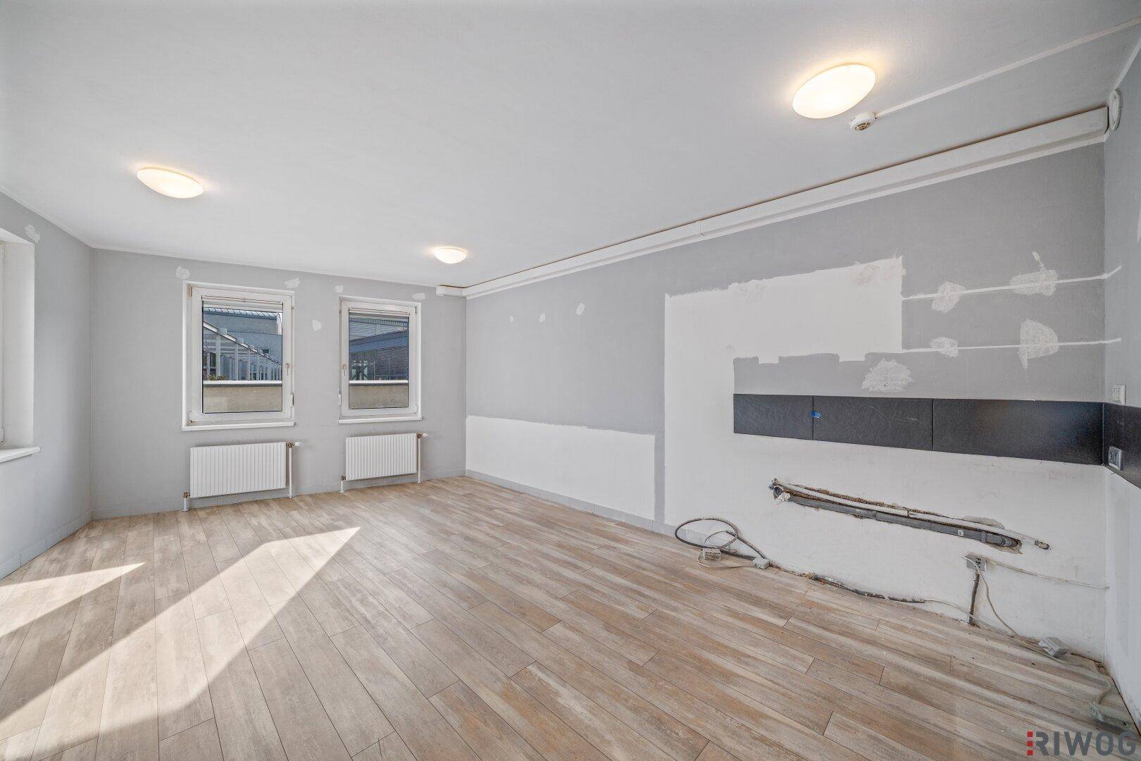Geräumiger Raum mit hellem Boden und vorbereiteter Wand für eine mögliche Küchenzeile oder Theke.
