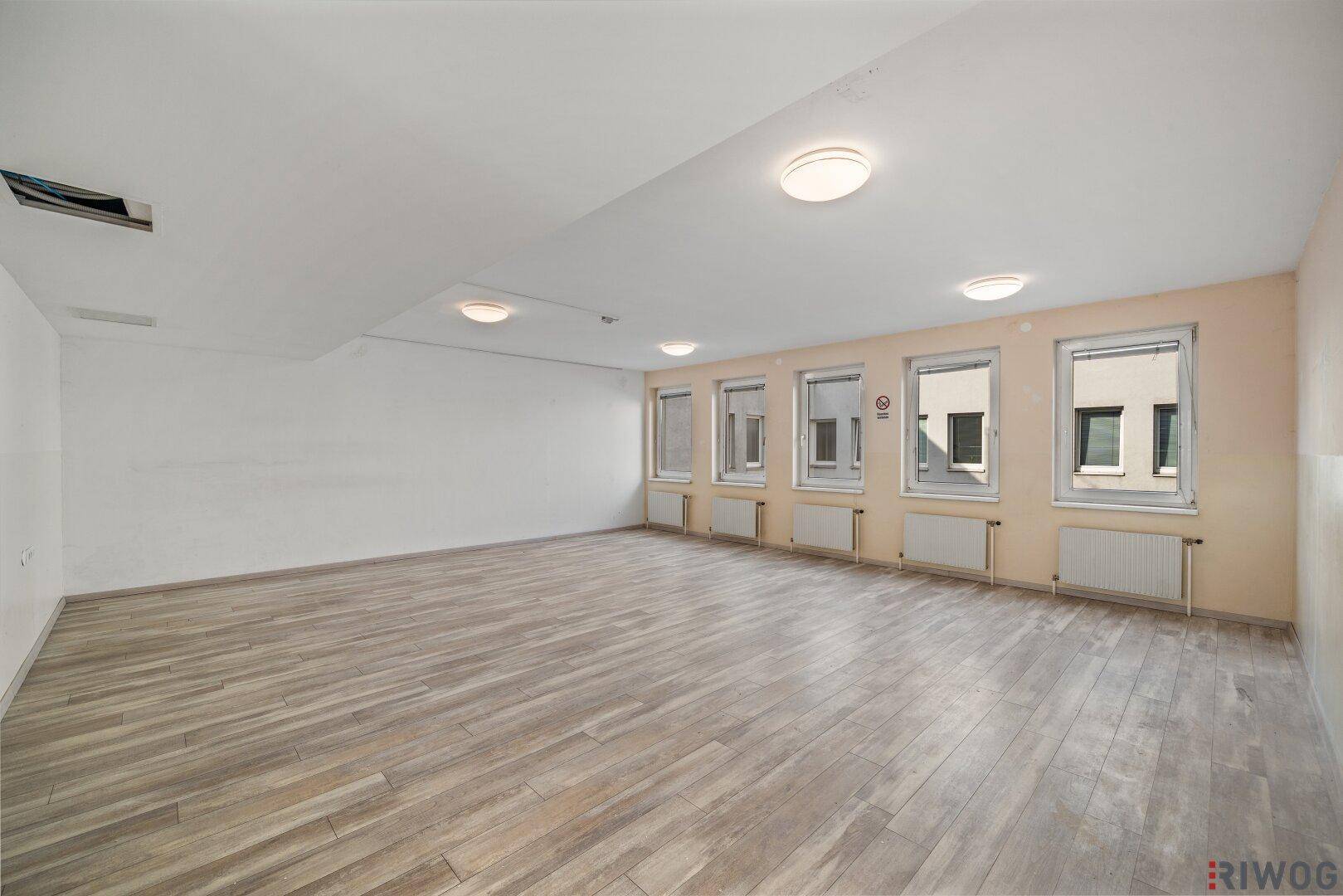 Leerer Gewerberaum mit hellem Boden und einer Fensterfront, die viel Tageslicht hereinlässt.
