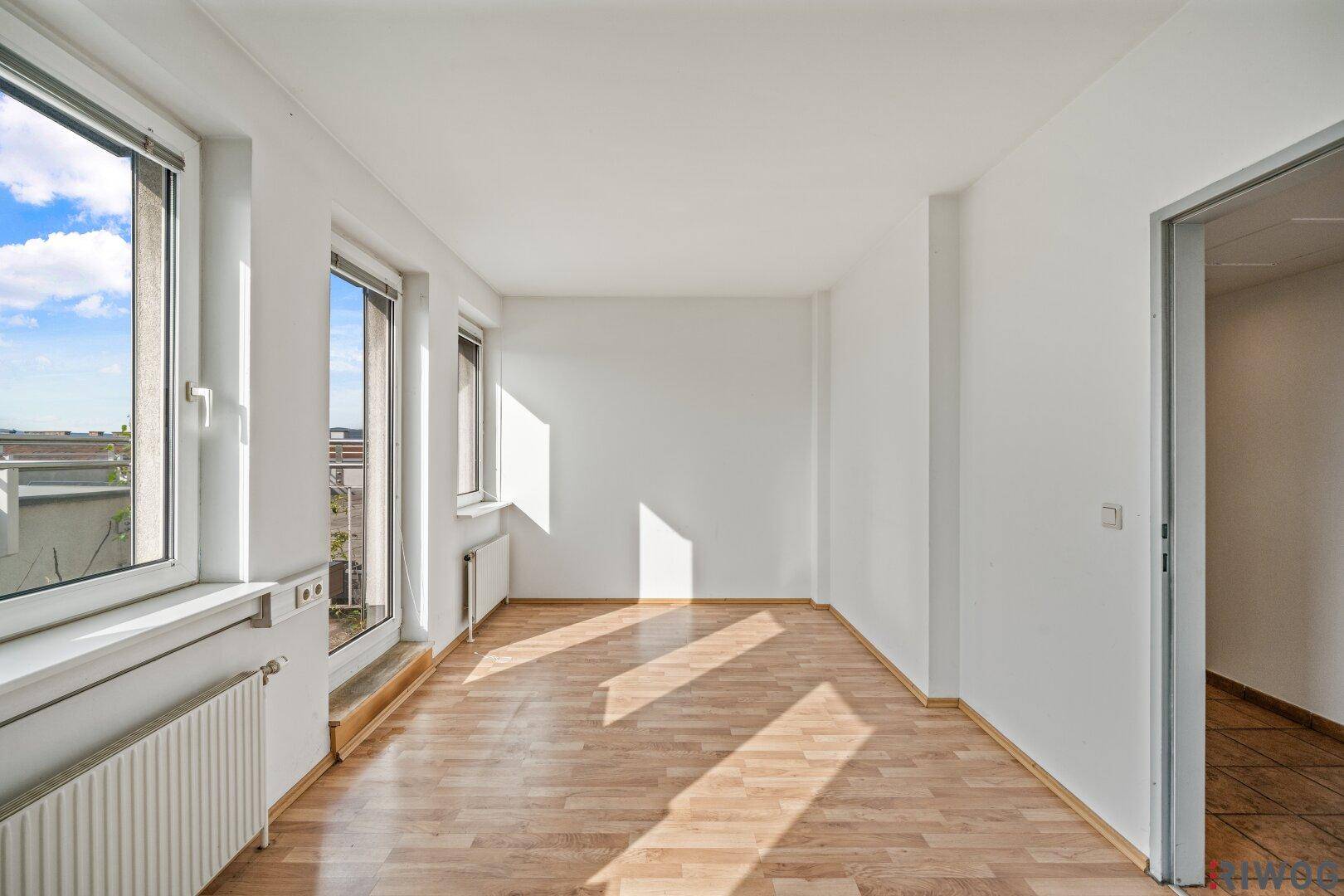 Heller, leerer Raum mit Laminatboden, großen Fenstern und Heizkörpern unter den Fensterbänken.