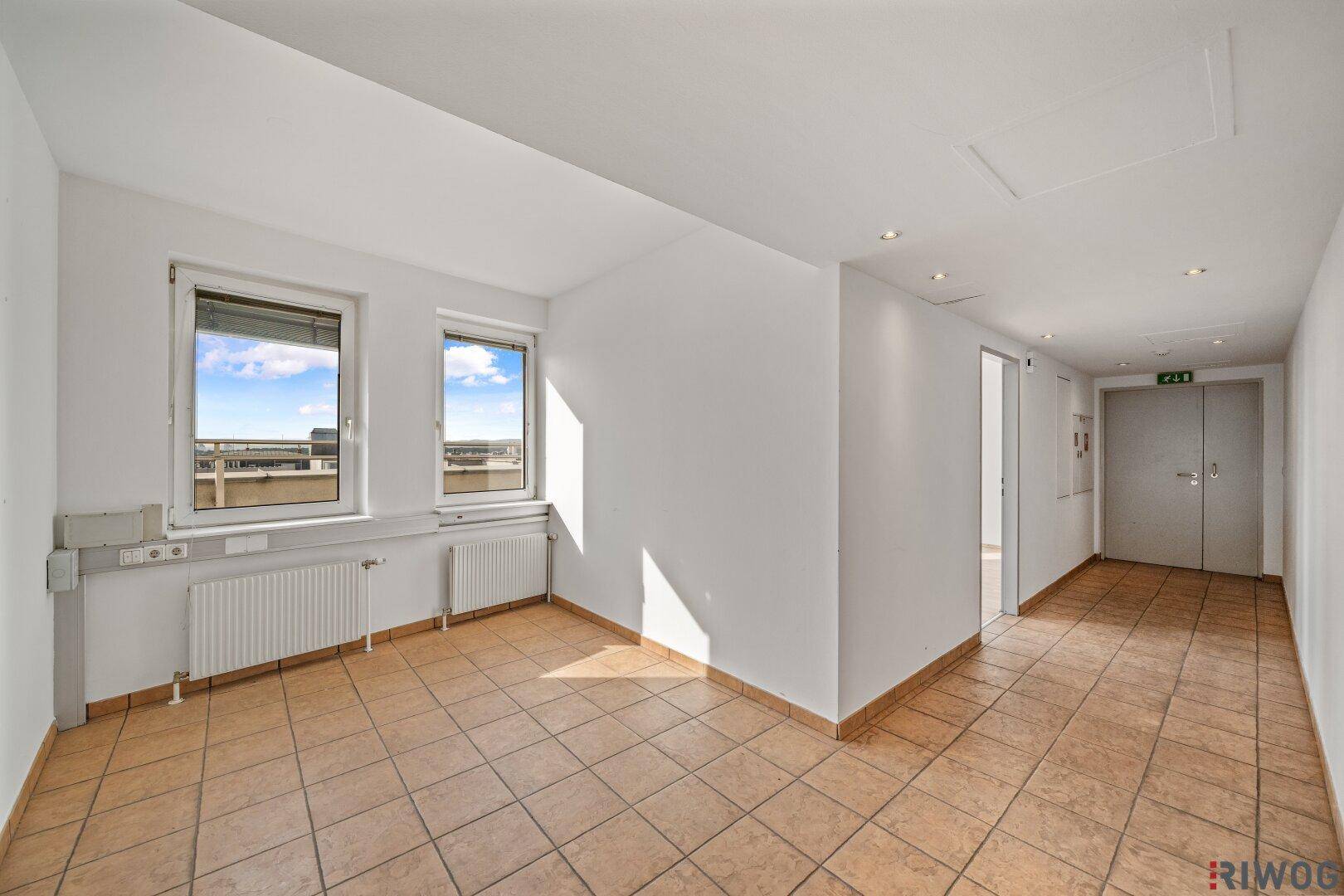 Geräumiger Raum mit hellbraunen Fliesen, Fenstern und Heizkörpern, ideal als Büro oder Gemeinschaftsbereich.