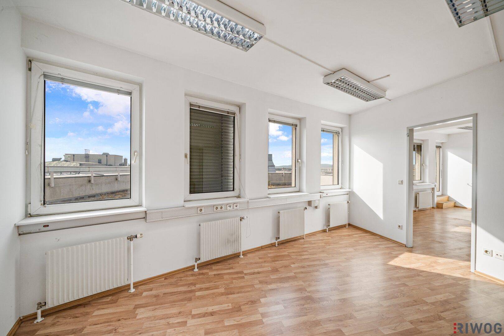 Heller Büroraum mit mehreren Fenstern, die viel Tageslicht hereinlassen, und einem Zugang zu einem weiteren Bereich.