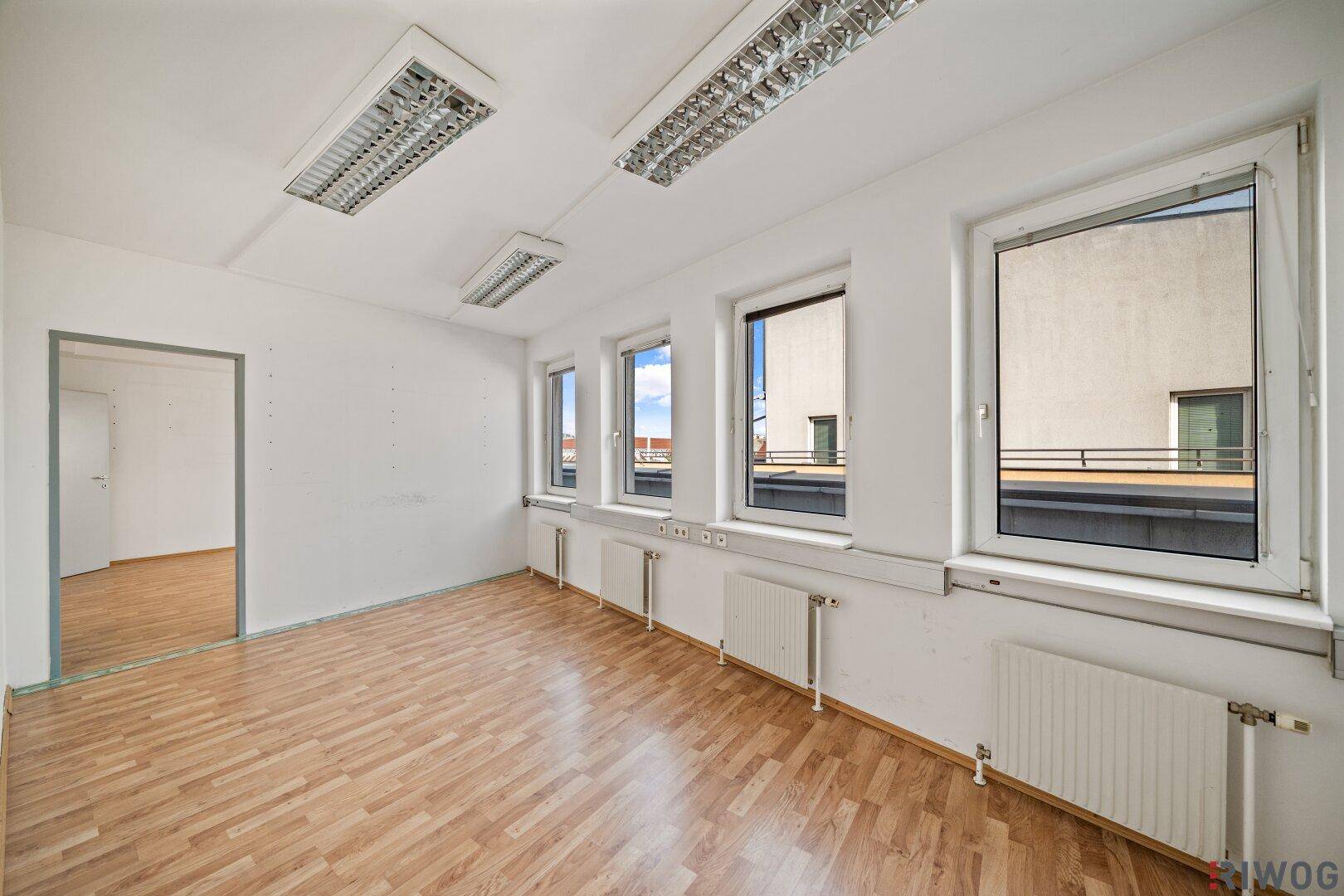 Leerer Büroraum mit Laminatboden, weißen Wänden und Blick aus den Fenstern auf ein Flachdach.