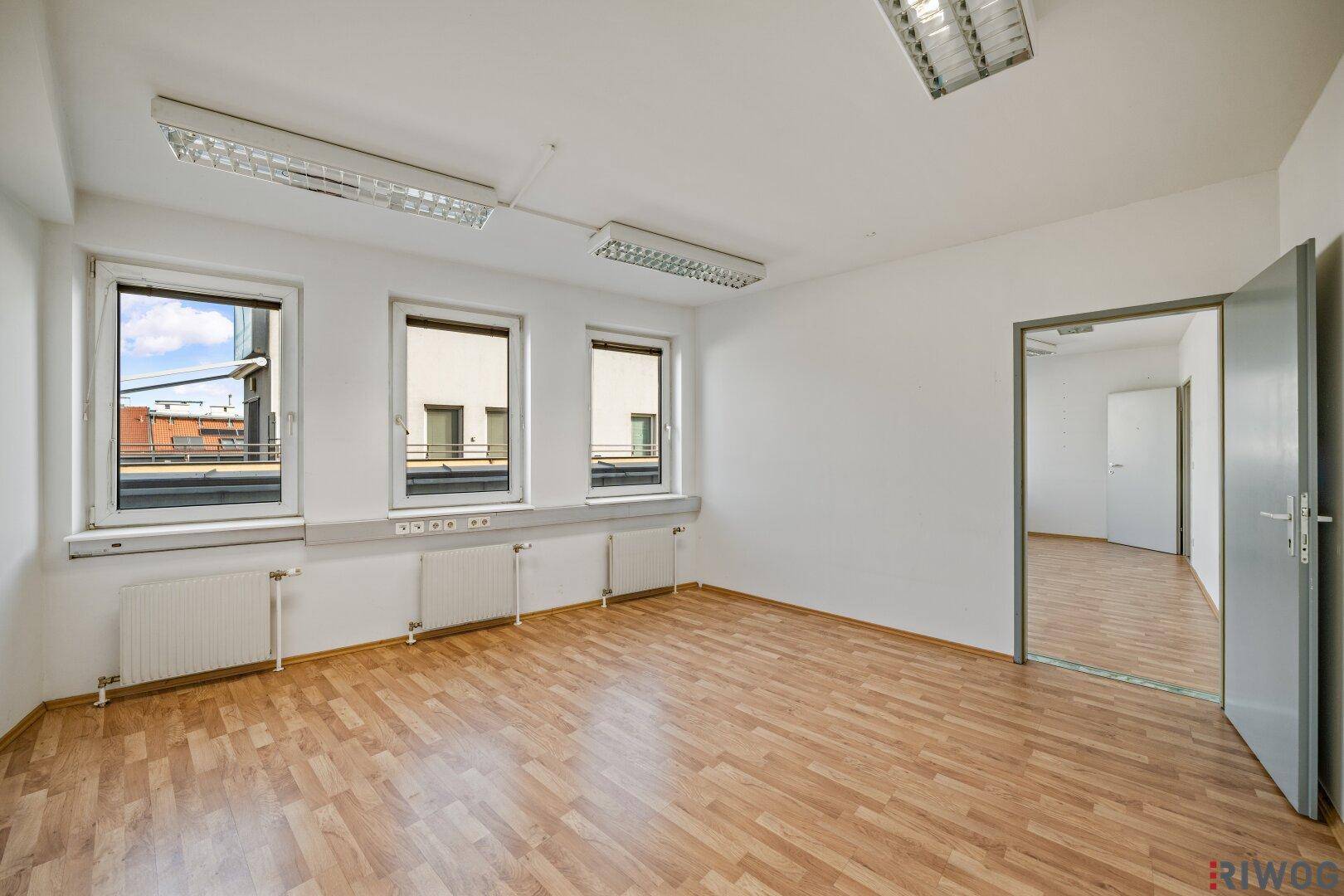 Helles, leeres Büro mit Laminatboden, Heizkörpern unter den Fenstern und Zugang zu einem weiteren Raum.
