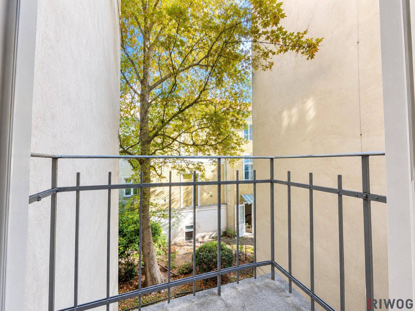 Kleiner Balkon mit schmiedeeisernem Geländer und Blick auf einen grünen Innenhof mit altem Baum.