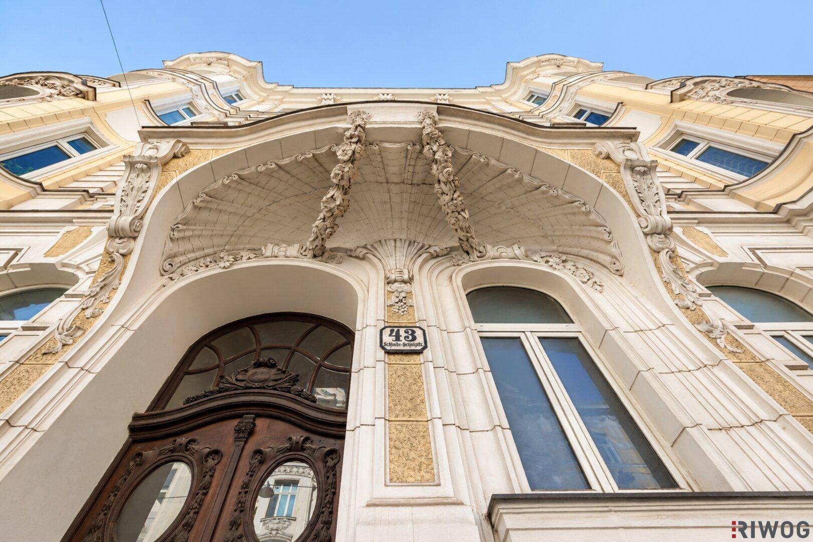 Blick nach oben auf das kunstvoll verzierte Eingangsportal eines historischen Gebäudes mit Stuckdetails.