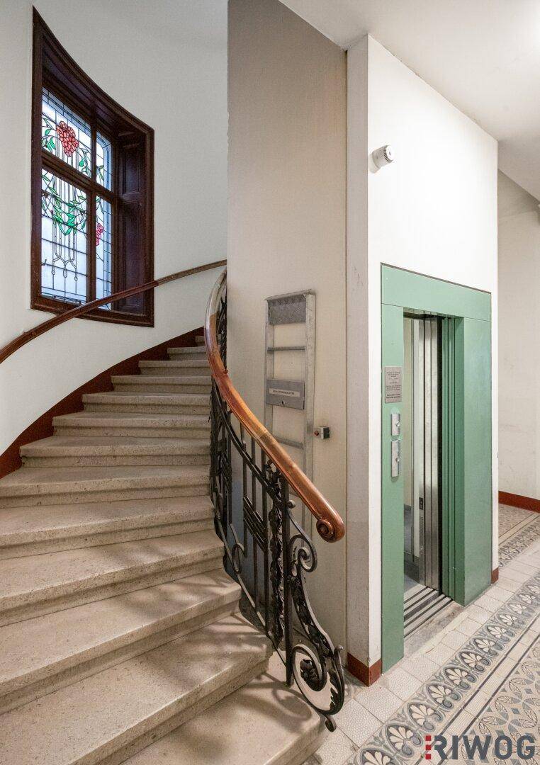 Historisches Treppenhaus mit Aufzug, verzierten Geländern und einem Buntglasfenster.