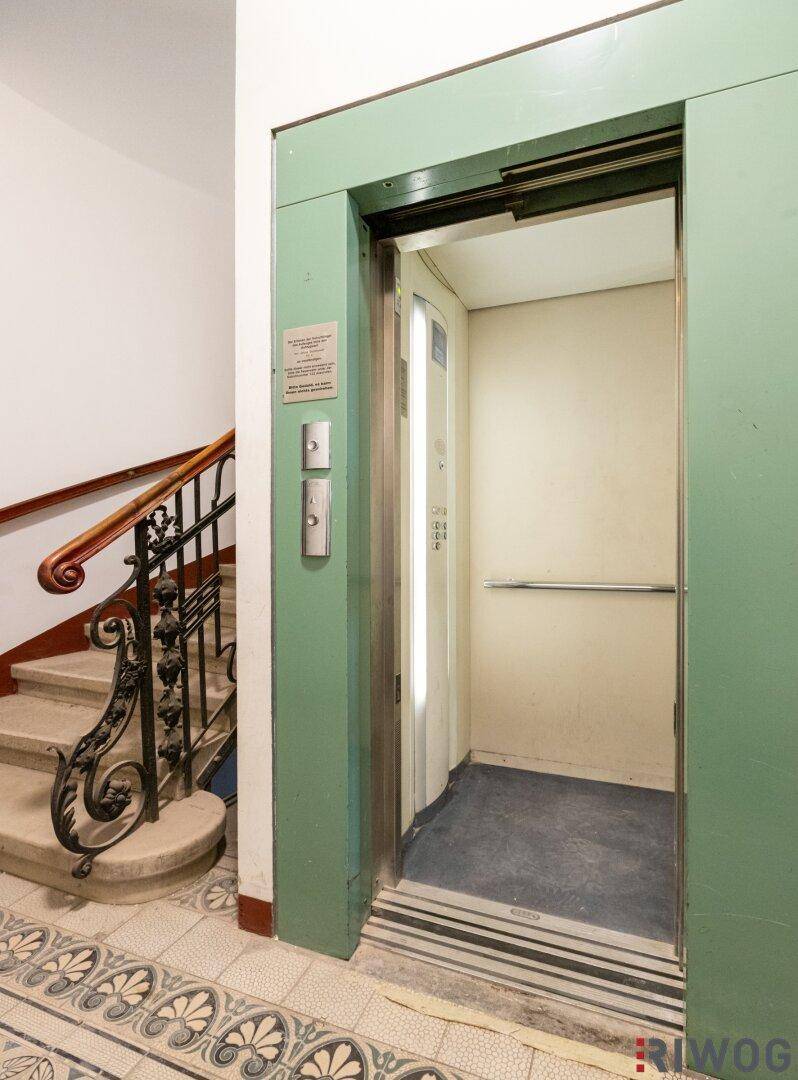 Detailansicht des Aufzugs im Treppenhaus mit grüner Tür und historischen Fliesenboden.