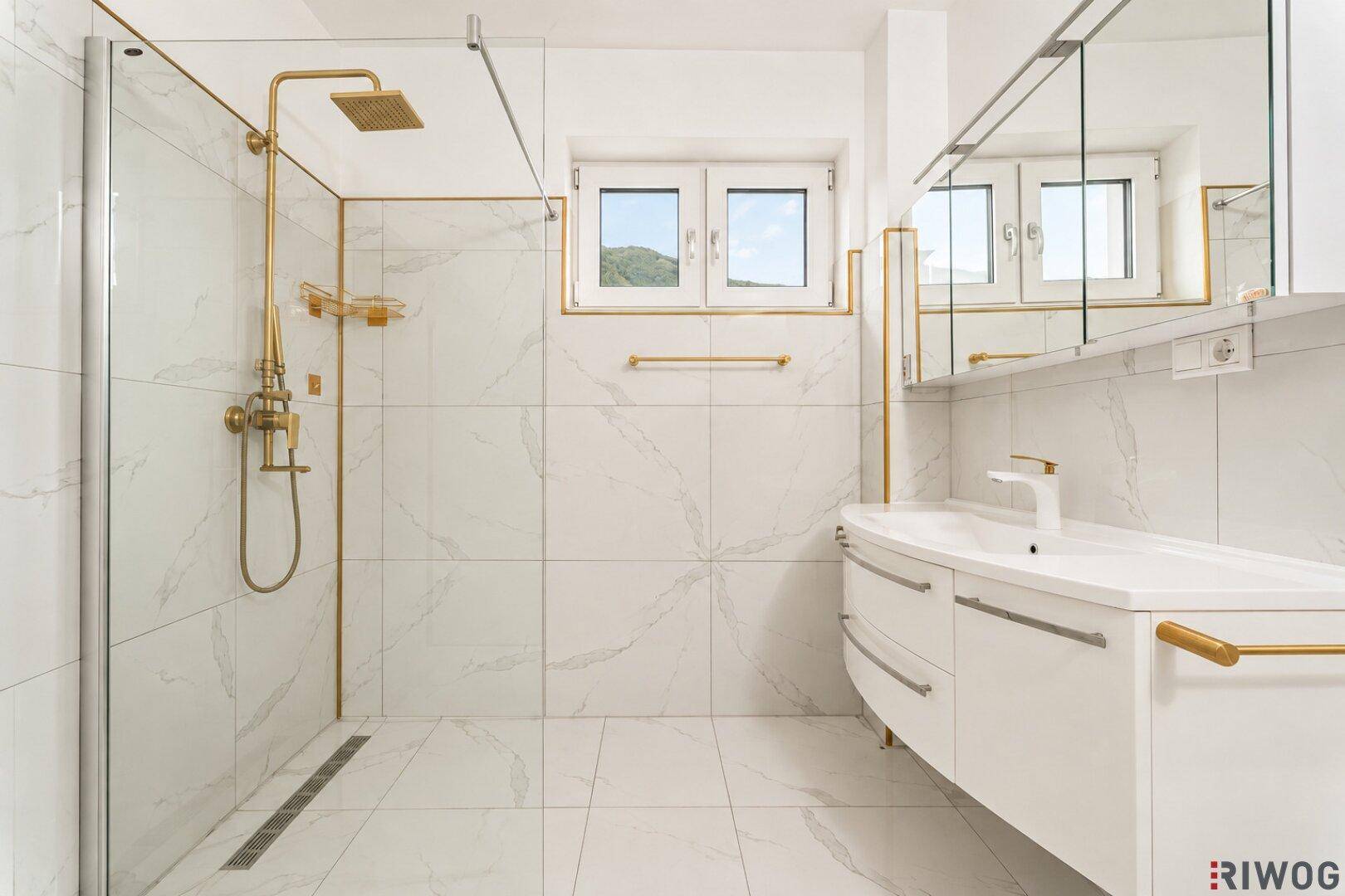 Modernes Badezimmer mit weißen Marmorfliesen, goldenen Armaturen, begehbarer Dusche und Fenster.