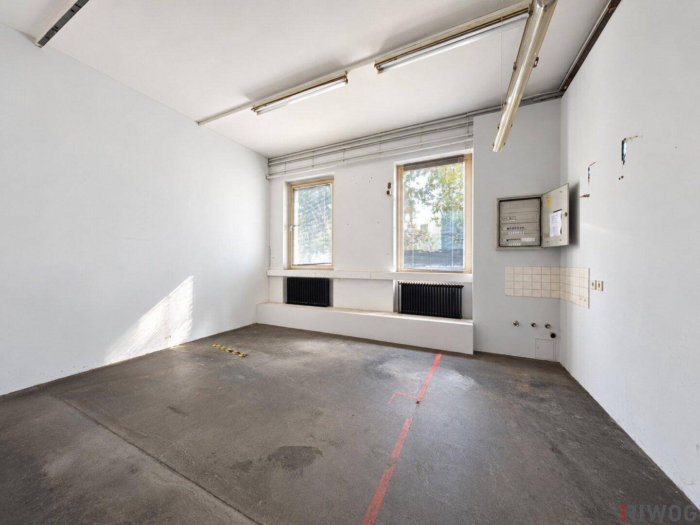 Kleiner Raum mit zwei Fenstern, Heizkörpern und einem Sicherungskasten an der Wand.