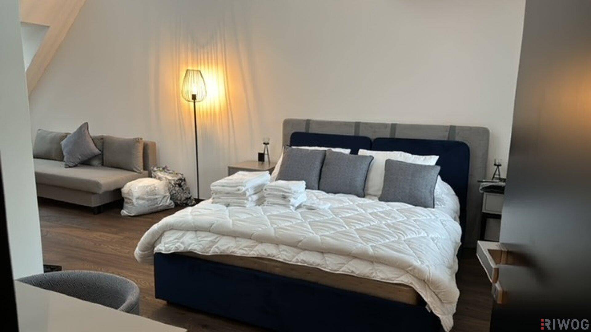 Helles Schlafzimmer mit Doppelbett, Sofa und stilvoller Stehlampe auf Holzboden.