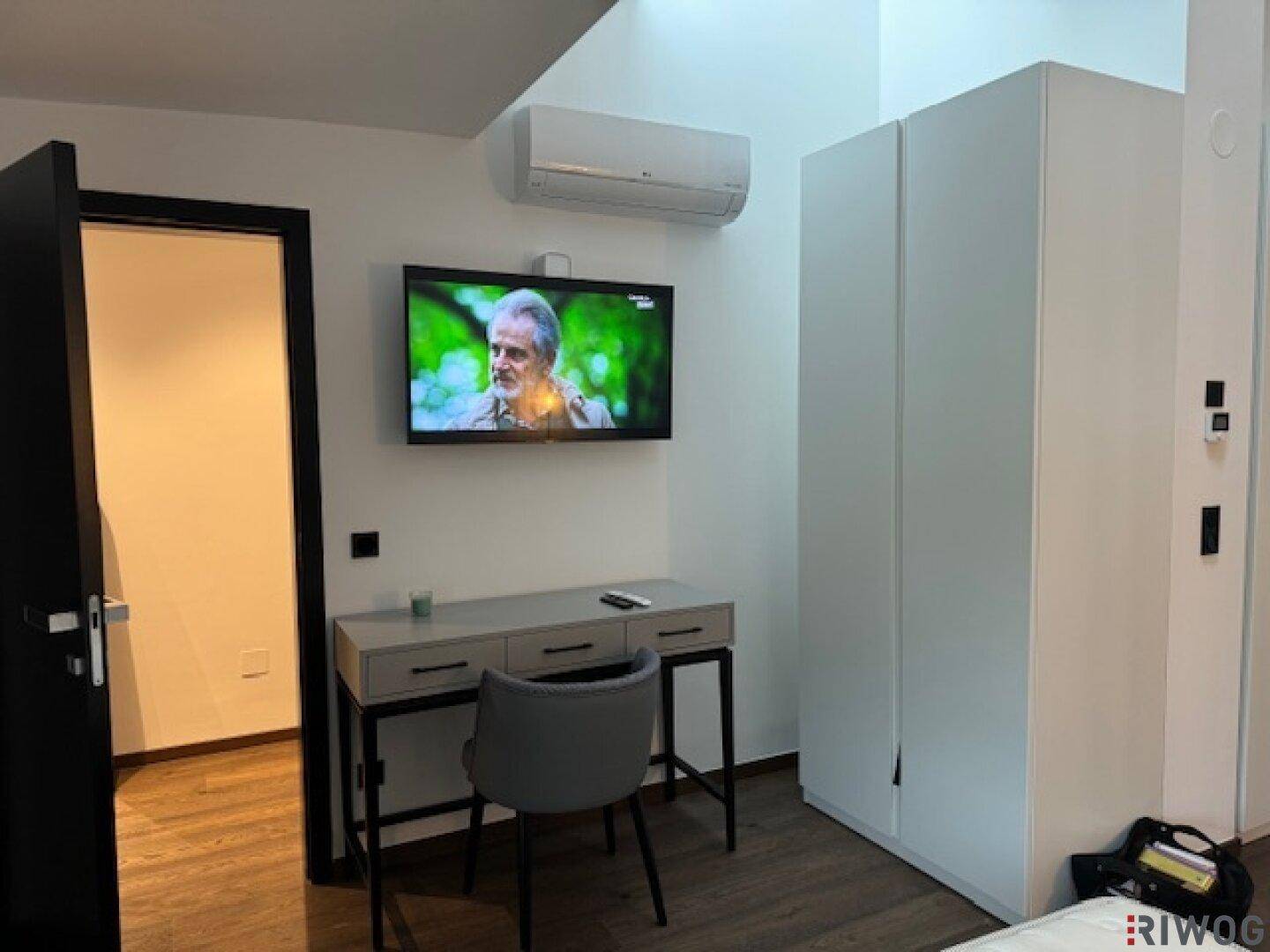 Schlafzimmer mit Schreibtisch, Wandfernseher, Klimaanlage und Einbauschrank.