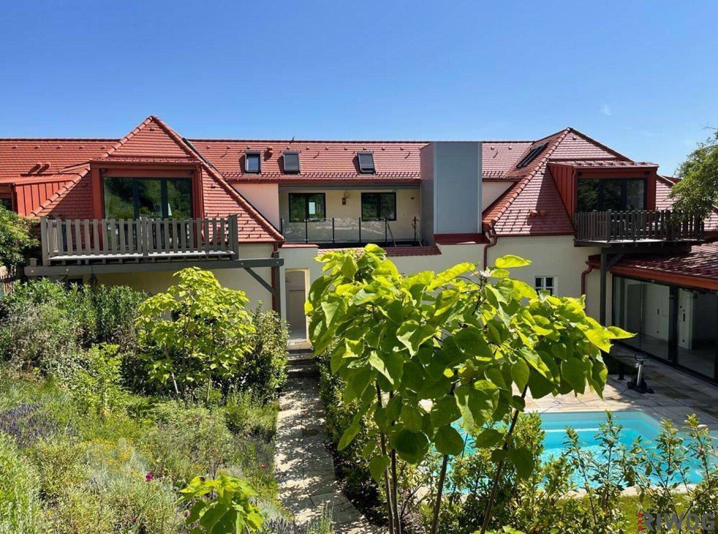 Gebäude mit rotem Mansarddach, Balkonen und einem gepflegten Garten mit Pool im Vordergrund.