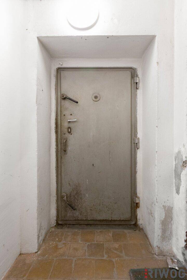Schwere, alte Stahltür in einem Kellerraum mit Fliesenboden und weiß gestrichenen Wänden, in gutem Zustand.