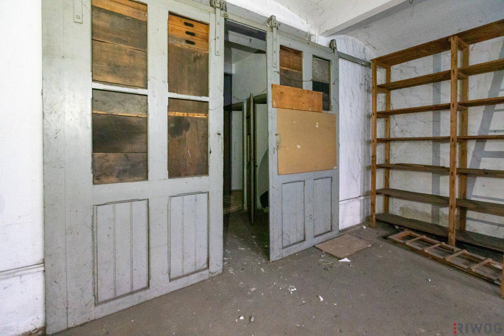 Alter Lagerraum mit großen Schiebetüren aus Holz und leeren Holzregalen an der Wand, renovierungsbedürftig.