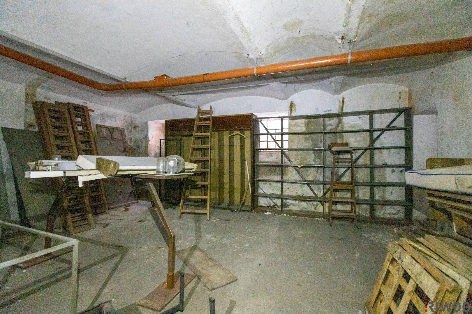 Unaufgeräumter Lagerraum mit alten Regalen, Holzpaletten und sichtbaren Rohren an der Decke.