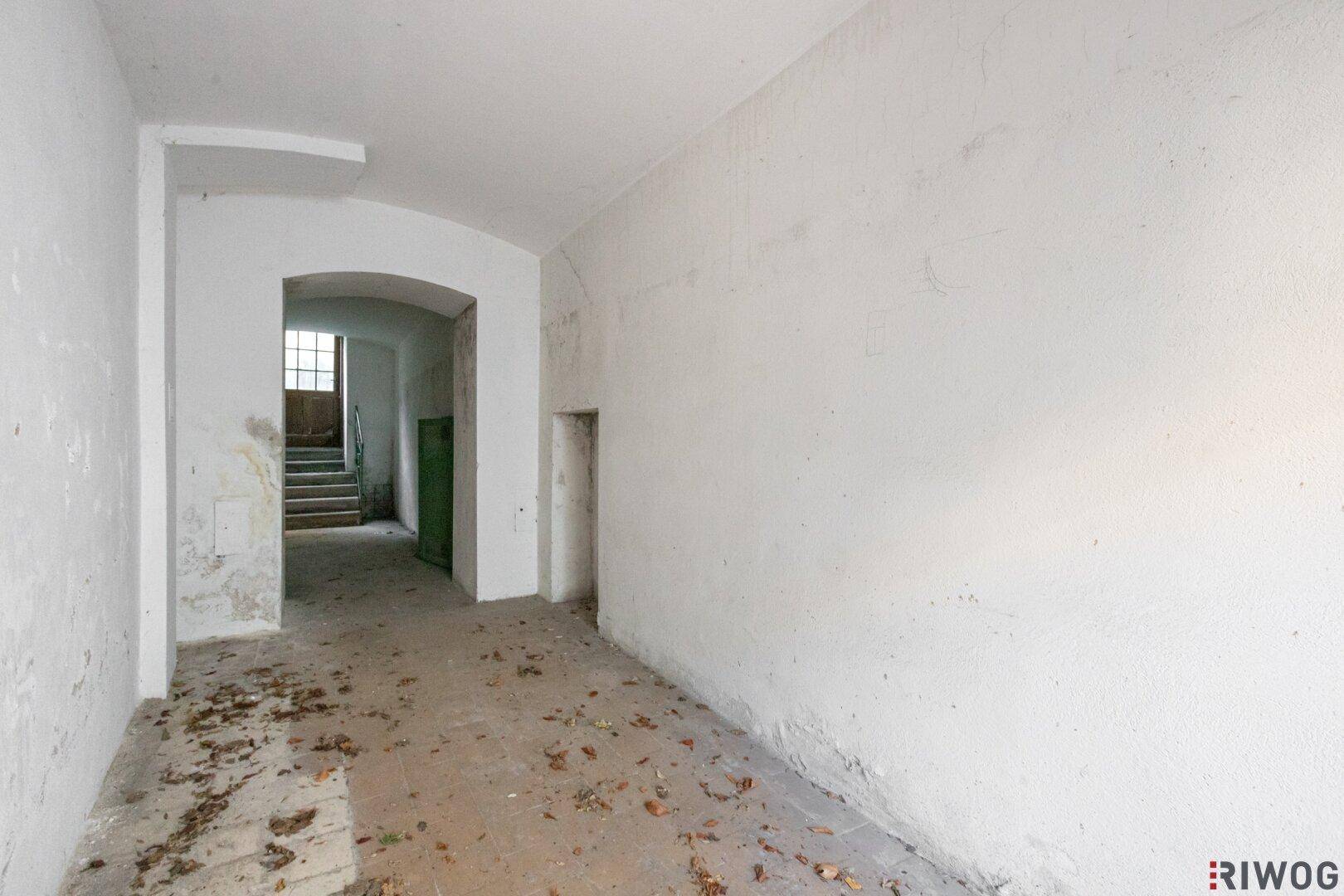 Langer, leerer Flur mit weiß gestrichenen Wänden und einem Treppenaufgang am Ende, in altem Zustand.