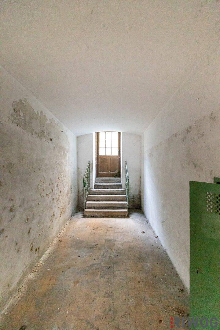 Langer Flur mit Treppe und alter Holztür am Ende, die ins Freie führt, mit natürlichem Licht.