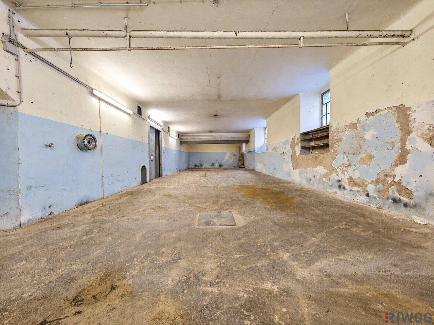 Weiterer Blick in den langen Lagerraum mit Betonboden, abblätternder Farbe an den Wänden und kleinen Fenstern.