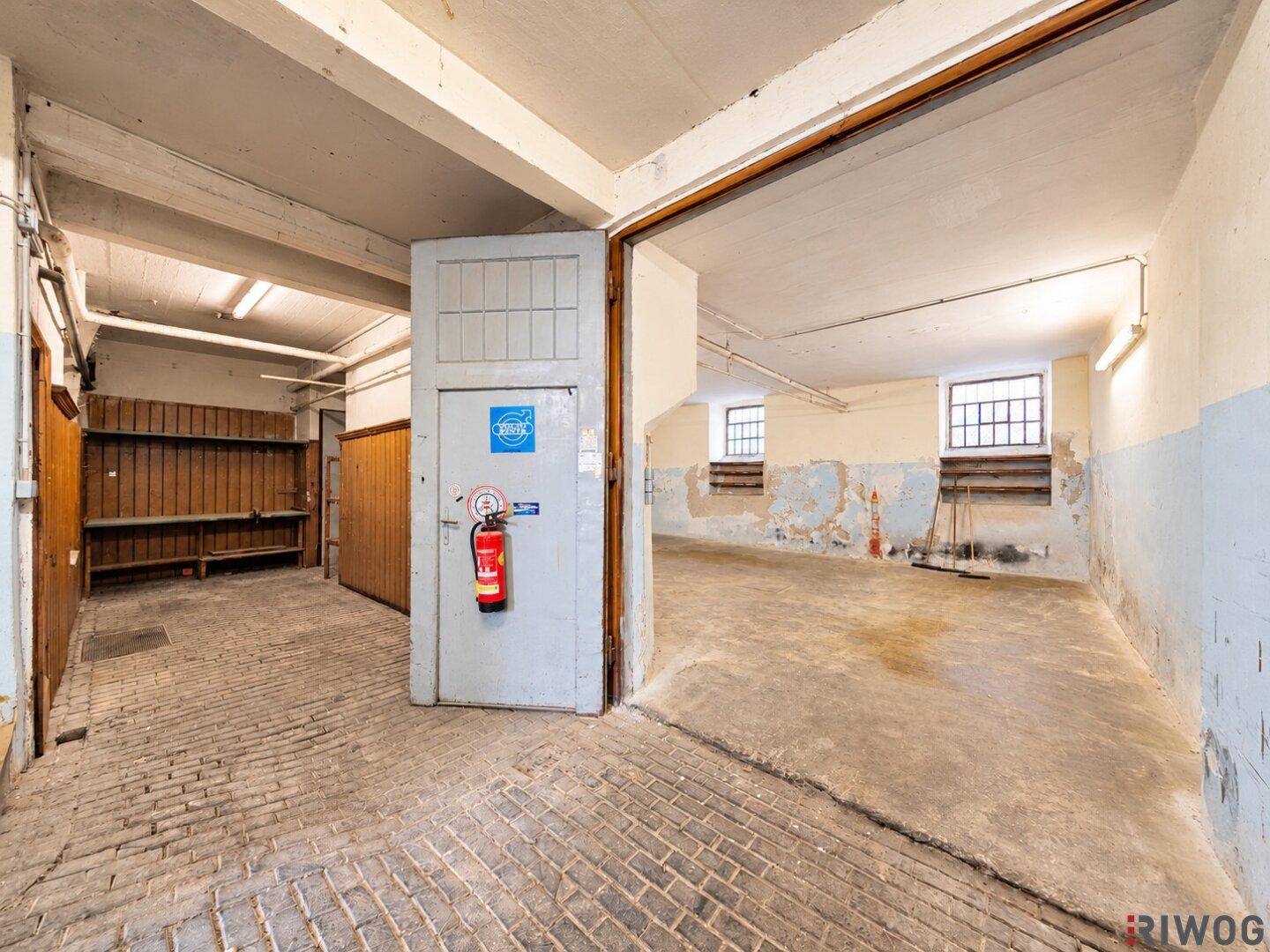 Übergangsbereich zwischen einem Lagerraum mit Holzverkleidung und einem größeren Bereich mit Betonboden und kleinen Fenstern.