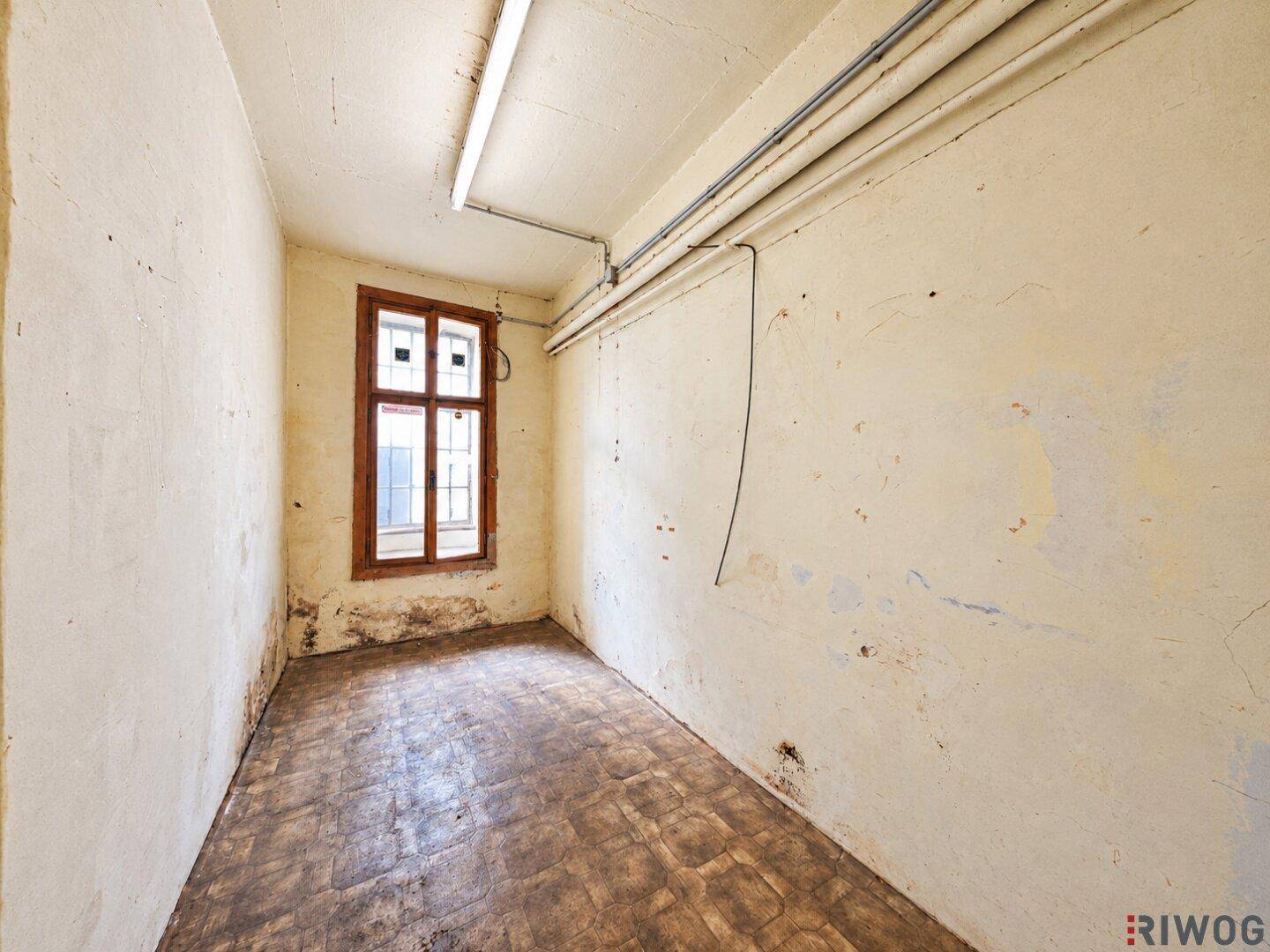 Kleiner, leerer Raum mit einem einzelnen Fenster, abblätternder Farbe an den Wänden und altem Bodenbelag.