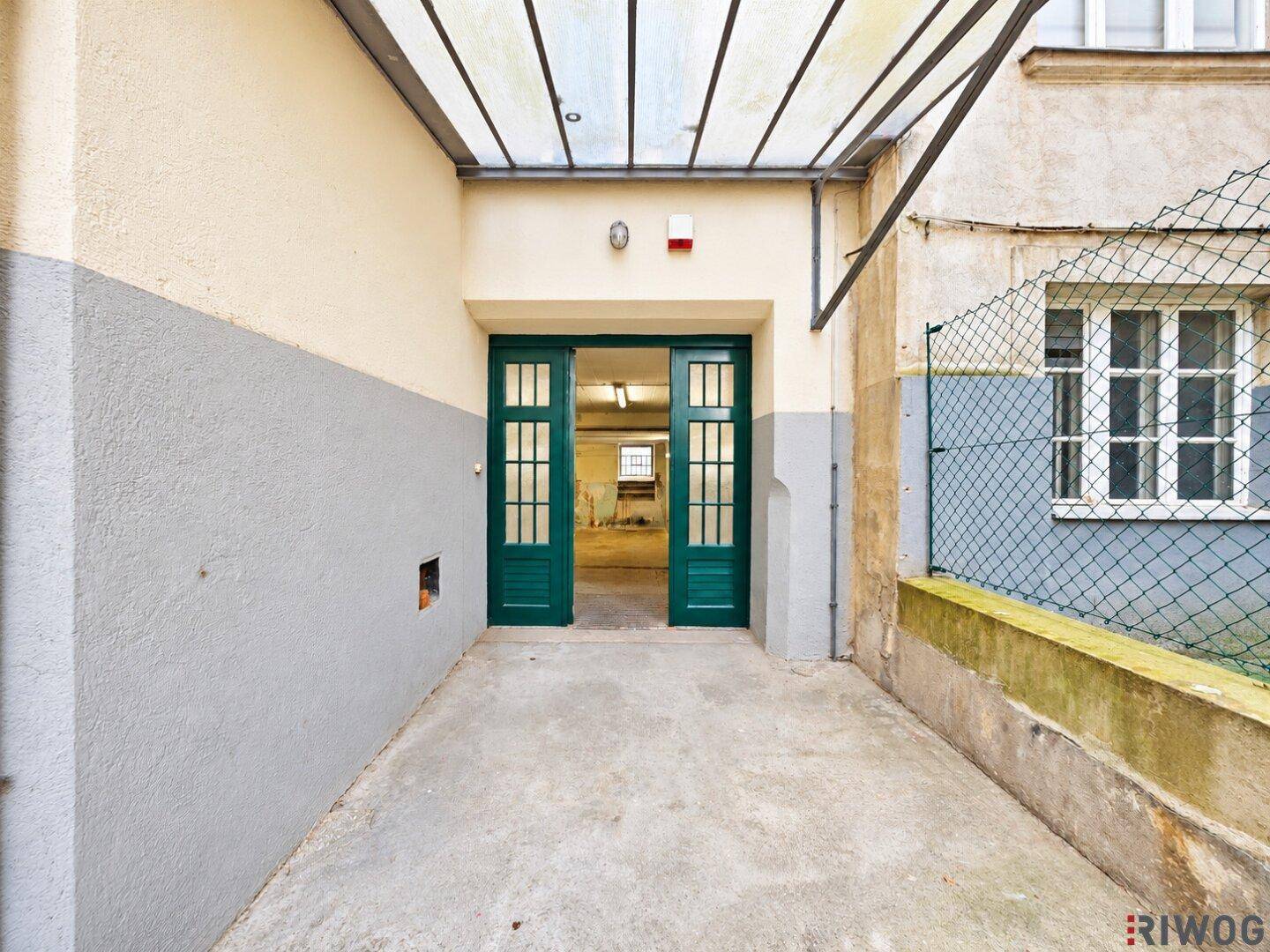 Außenansicht eines Eingangs mit grünen Doppeltüren, überdacht von einem Glasvordach, und einer Betonrampe.