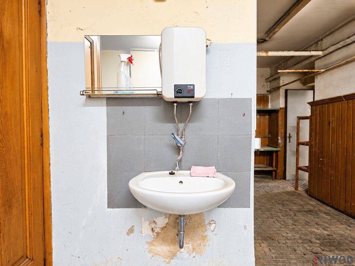 Nahaufnahme eines Waschbeckens mit Warmwasserbereiter über einem gefliesten Wandbereich, im Hintergrund Holzverkleidung.