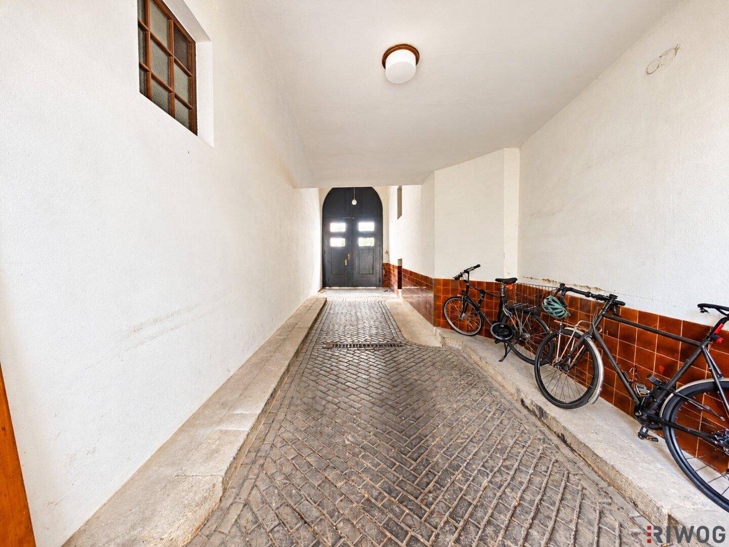 Langer, schmaler Durchgang mit gepflasterter Rampe und Fahrrädern an der Seite, führt zu einer dunklen Doppeltür.