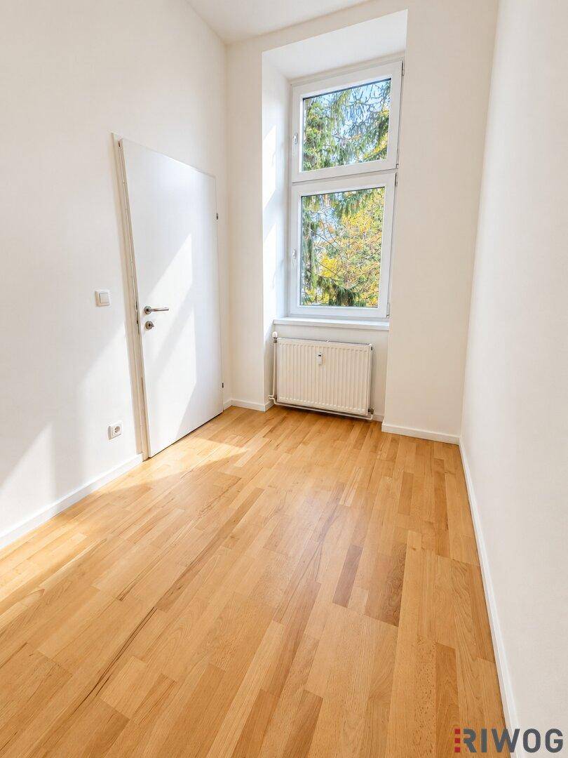 Helles, leeres Zimmer mit Parkettboden und einem hohen Fenster.