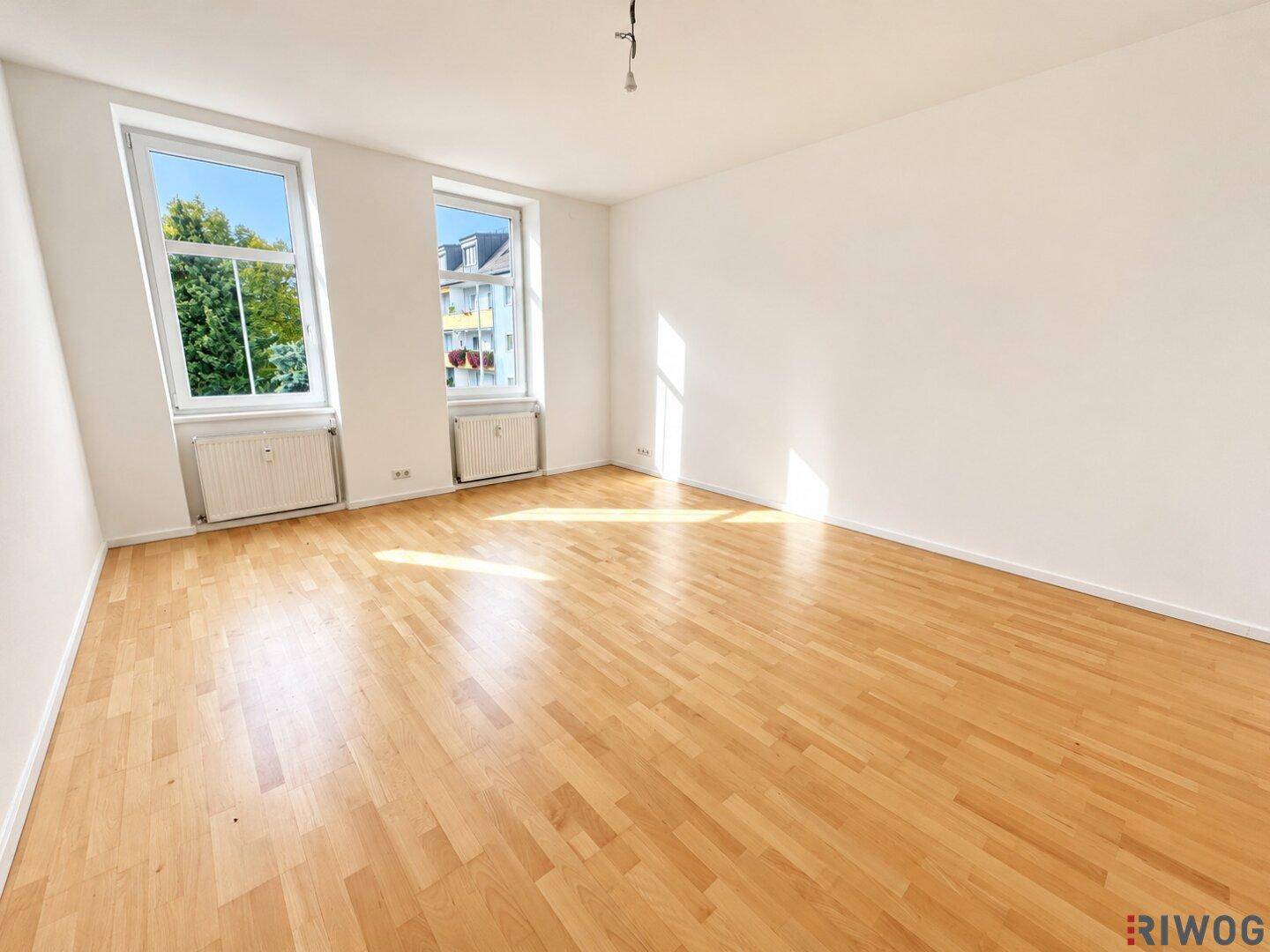 Geräumiges, leeres Wohnzimmer mit hellem Parkettboden und zwei großen Fenstern.