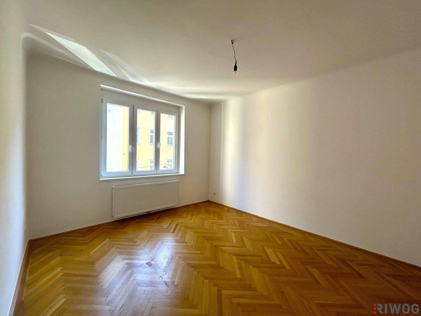 Geräumiges Zimmer mit klassischem Fischgrätparkett und Blick auf die gegenüberliegende Gebäudefassade.