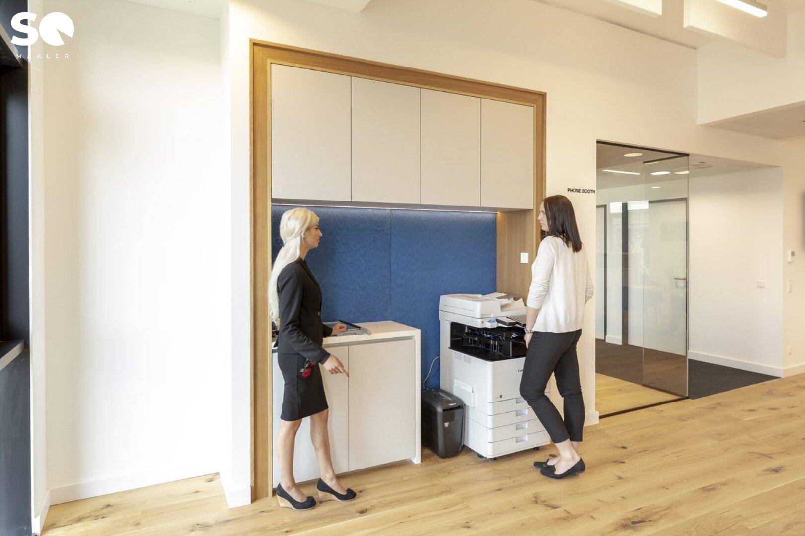 Funktionaler Bereich mit Drucker und Stauraum, umgeben von weißen Wänden und Holzboden.