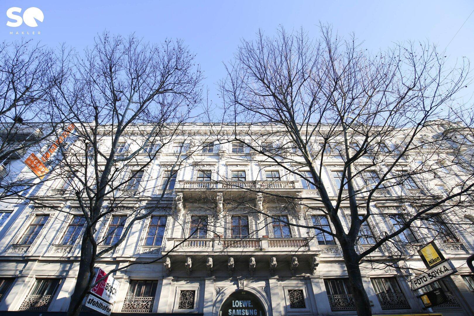 Historische Fassade eines Gebäudes mit Balkonen und kahlen Bäumen im Vordergrund, unter blauem Himmel.