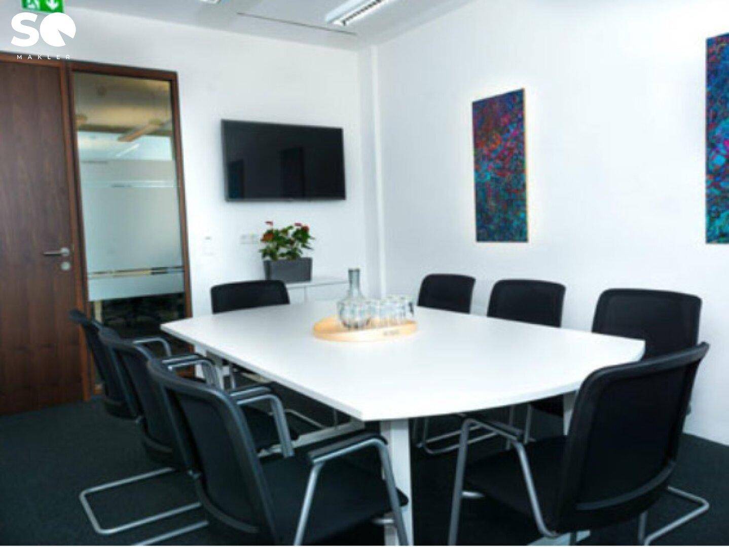 Konferenzraum mit großem weißen Tisch, bequemen Stühlen und Wandbildschirm für Präsentationen.