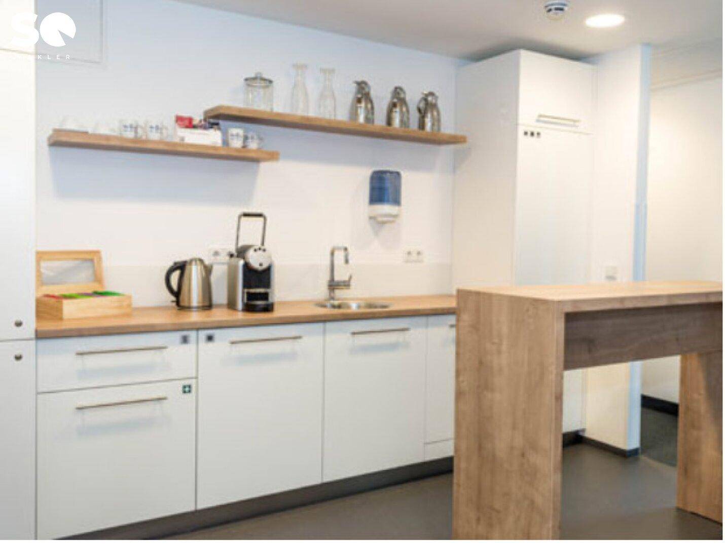 Moderne Teeküche mit weißen Schränken, Holztheke und integrierter Kaffeemaschine.