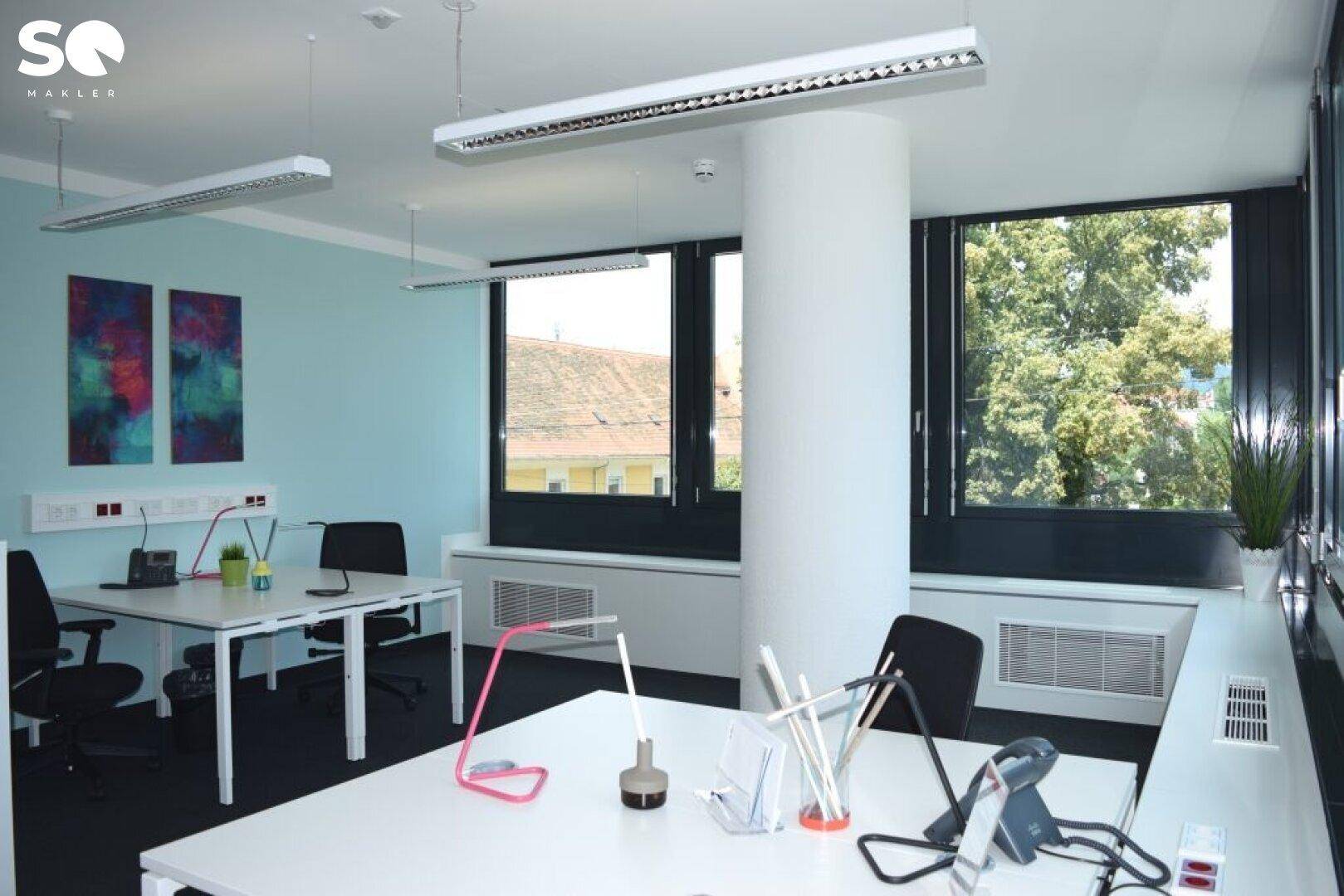 Helles Büro mit blauen Wänden, großen Fenstern und Blick ins Grüne, ausgestattet mit modernen Schreibtischen.