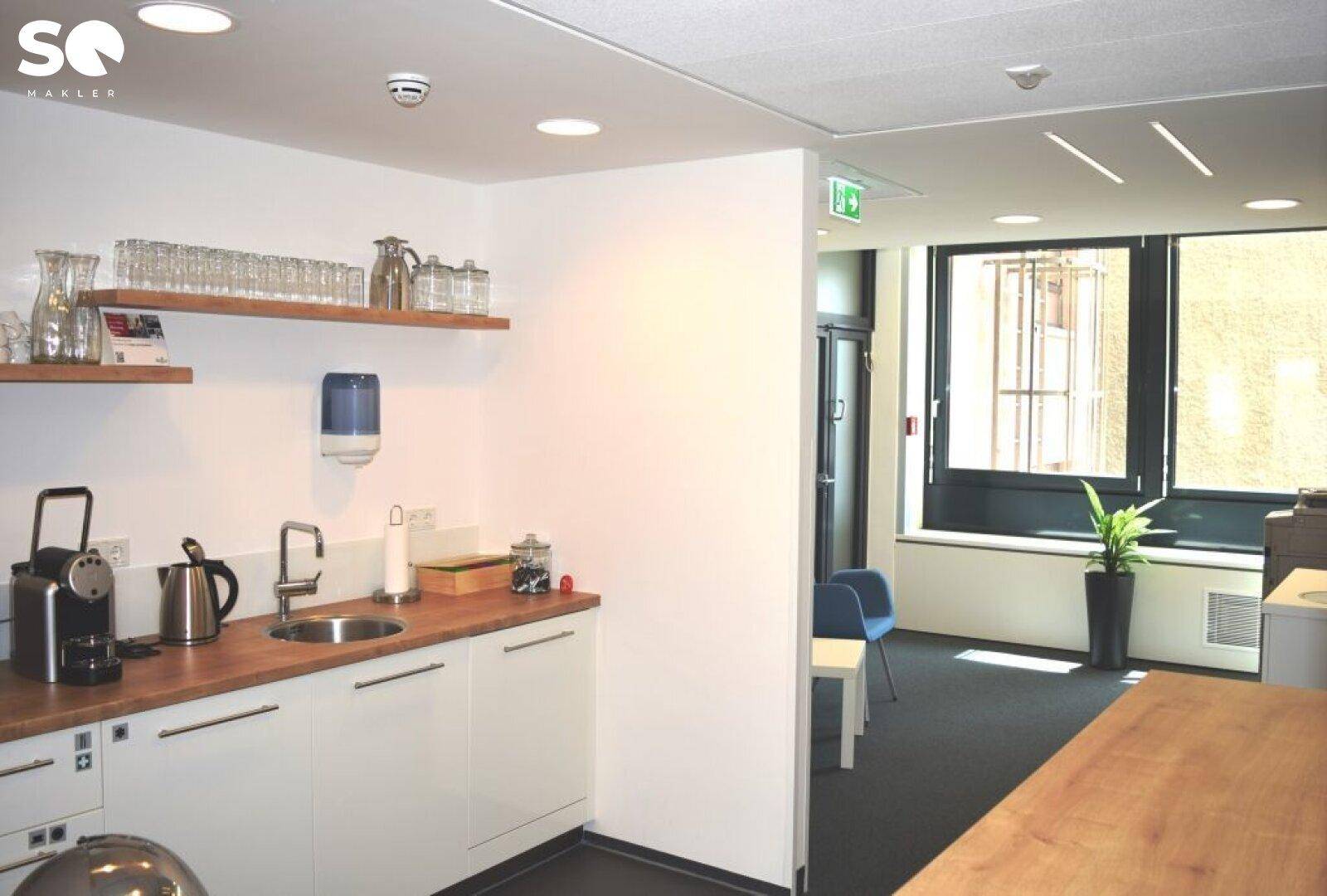 Kleine, funktionale Teeküche mit Spüle, Holztheke und Regalen für Geschirr in einem Bürogebäude.