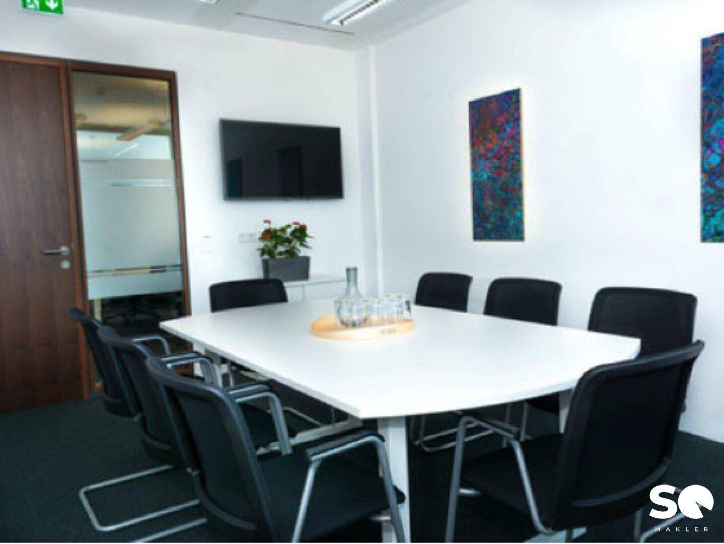 Konferenzraum mit großem weißen Tisch, bequemen Stühlen und Wandfernseher für Präsentationen.